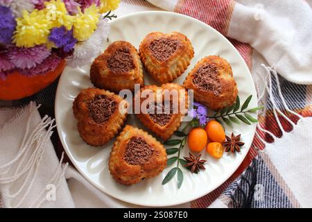 Köstliche Muffins in Herzform mit Schokoladenfüllung sind wunderschön auf einem weißen Teller angeordnet, begleitet von Kumquats, Sternanis und violetten Blüten Stockfoto