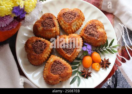 Köstliche herzförmige Muffins gefüllt mit Schokolade werden auf einem weißen Teller serviert, garniert mit Kumquat und Sternanis, was eine gemütliche Herbstszene erzeugt Stockfoto