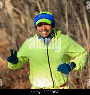 Smithtown, New York, USA - 14. Dezember 2024: Ein Mann in heller Laufausrüstung lächelt und läuft an einem sonnigen Tag auf einem Waldweg, der Natur und Natur umschließt Stockfoto