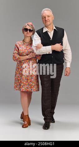 Paar Geharm im Arm, Frau in hellem Kleid und offenen Schuhen, Mann in dunkler karierter Hose und schwarzer Weste vor grauem Hintergrund. Stockfoto