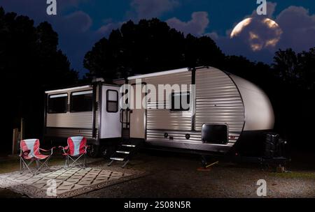 Mond, der hinter einem dunklen Wald aufsteigt, der einen beleuchteten Campingwagen umgibt, der für die Nacht aufgestellt ist. Stockfoto