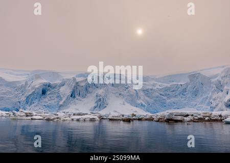 Eine niedrige Sonne scheint schwach in einem nebeligen Himmel über einer verschneiten Landschaft aus Gletschern und Spalten der antarktischen Küste am Portal Point Stockfoto