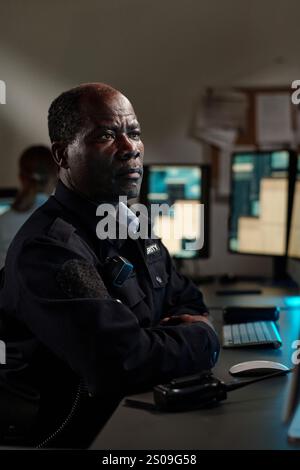 Reifer männlicher Sicherheitsbeamter in schwarzer Uniform, der am Schreibtisch im Kontrollraum sitzt und auf den Bildschirm des Desktop-Computers blickt Stockfoto