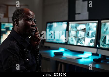 Seriöser, ausgereifter Sicherheitsbeamter mit Telefonempfänger am Ohr, der am Arbeitsplatz vor cctv-Videos auf Computerbildschirmen sitzt Stockfoto