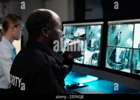 Ernster reifer Offizier in Uniform, der eine Tasse Kaffee trinkt, während er vor dem Computerbildschirm sitzt und cctv-Videos ansieht Stockfoto