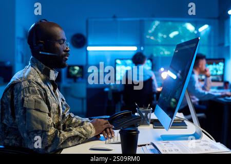 Junger afroamerikanischer Militärangehöriger in Headset, Brille und Tarnuniform, der auf den Desktop-Bildschirm schaut Stockfoto