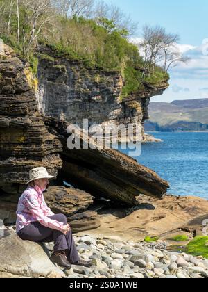 Erwachsene Frau mit Sonnenhut, die an einem sonnigen Tag im Mai an einem felsigen Strand sitzt, Glasnakille bei Elgol, Isle of Skye, Schottland, Großbritannien Stockfoto