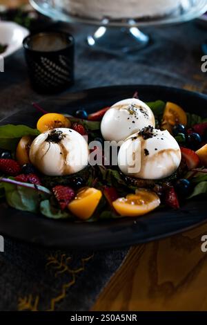 Ein Gericht Burrata-Käse wird auf frischem Gemüse serviert, umgeben von Erdbeeren, Heidelbeeren und gelben Tomaten, mit Balsamico-Glasur auf einem dunklen Ta Stockfoto
