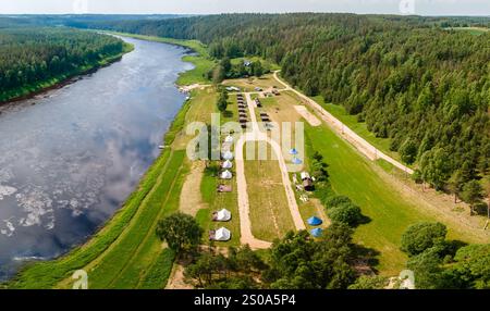 Ein sich windender Fluss fließt durch einen Campingplatz mit Zelten und kleinen Bauten. Weitläufige grüne Felder und dichte Wälder umgeben das Flussgebiet. Stockfoto
