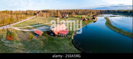 In der Nähe eines gewundenen Wasserwegs befindet sich eine Ansammlung von Gebäuden mit rotem Dach, umgeben von offenen Feldern und einem dichten Wald von blattlosen Bäumen unter klarem Himmel. Stockfoto