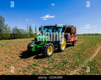 Ein grüner Traktor zieht einen roten Anhänger mit Heuballen über ein gepflügtes Feld. Bäume umranden das Feld, und helles Sonnenlicht unterstreicht die lebhafte Atmosphäre Stockfoto