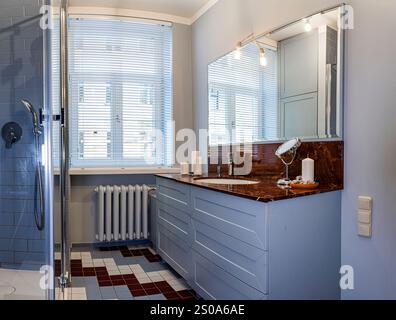 Das moderne Badezimmer verfügt über einen Waschtisch mit Marmorplatte, weiche blaue Schränke und einen geometrischen Fliesenboden. Natürliches Licht filtert durch ein Fenster mit Sonnenblende Stockfoto
