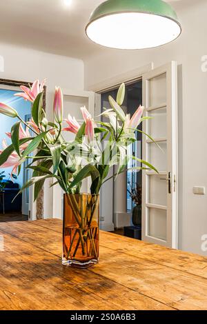 Ein Strauß rosa Lilien sitzt in einer braunen Glasvase auf einem Holztisch. Weiße Glastüren öffnen sich zu einem Raum mit dunkelblauem Stuhl und grünem Pendelleuchte Stockfoto