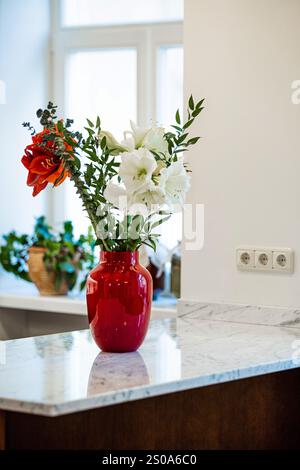 Eine leuchtend rote Vase mit weißen und roten Blumen steht auf einer Marmorarbeitsfläche. Natürliches Licht aus einem Fenster beleuchtet die Szene, mit einer Topfpflanze auf t Stockfoto