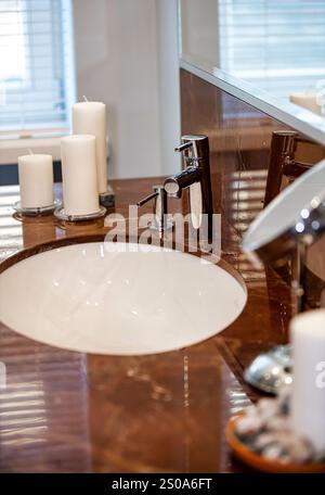 Das moderne Waschbecken im Bad verfügt über eine polierte Ablagefläche aus braunem Marmor, einen eleganten Wasserhahn und weiße Kerzen. Ein runder Spiegel reflektiert weiches Licht von wi Stockfoto