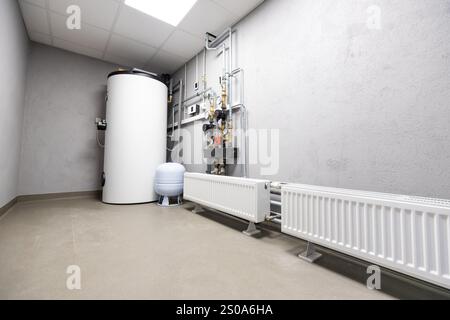 Innenansicht eines Versorgungsraums mit großem Weißwasserbereiter und blauem Drucktank. Ordentlich organisierte Rohre und Ventile säumen die gefliesten Wände. Stockfoto