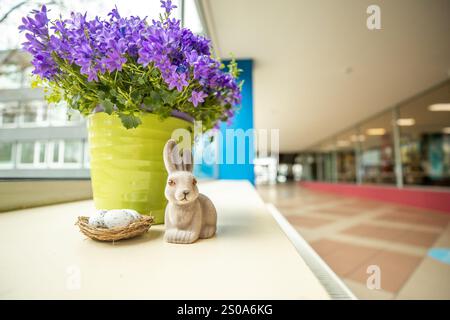 Ein hellgrüner Topf mit violetten Blüten sitzt auf einem Felsvorsprung, begleitet von einem Keramikkaninchen und einem Nest mit gesprenkelten Eiern vor einem modernen Interio Stockfoto