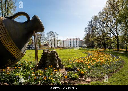 Ein großes Gebäude befindet sich vor einem Garten mit blühenden Tulpen und einer großen dekorativen Urne. Hohe Bäume umrahmen die Szene unter einem klaren blauen Himmel. Stockfoto