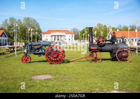 Zwei Vintage-Traktoren mit roten Rädern sitzen auf Gras, mit einem großen Gebäude mit einem roten Dach und einer weißen Fassade im Hintergrund unter einem klaren blauen Sch Stockfoto