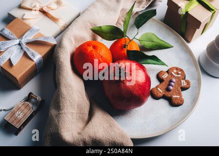 Festliches Stillleben mit Granatapfel, Orangen, Lebkuchenkeksen und gewickelten Geschenken auf einem weißen Teller. Weihnachtskonzept. Draufsicht. Stockfoto