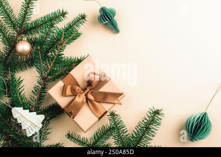 Weihnachtsliege mit grünen Tannenzweigen, einem gewickelten Geschenk mit brauner Schleife und Papierornamenten auf beigefarbenem Hintergrund. Draufsicht, Kopierraum. Stockfoto