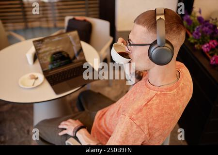 Hochwinkelaufnahme eines männlichen IT-Programmierers in kabellosen Kopfhörern, der Kaffee trinkt, während er beim Arbeiten am Tisch im Außenbereich des gemütlichen Cafés, Kopierraum, Kaffee schreibt Stockfoto