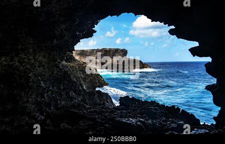 Eine malerische Aussicht auf das Meer durch eine felsige Höhlenöffnung, die Wellen zeigt, die gegen eine Klippe in der Ferne unter einem hellblauen Himmel mit flauschigem Himmel krachen Stockfoto