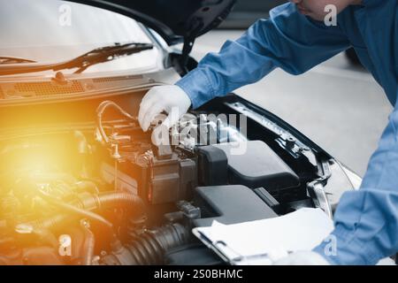 Fokussierter asiatischer Mechaniker mit einem Klemmbrett, der den Motor in der Garage überprüft. Expertise im Autoservice Stockfoto