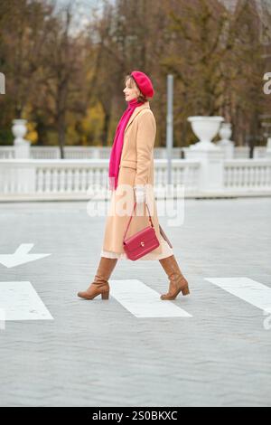 Eine Frau mit beigefarbenem Mantel, roter Baskenmütze und hellem Schal spaziert über einen Quersteg in einer malerischen Parklandschaft und präsentiert elegante Mode und Herbstszenen Stockfoto