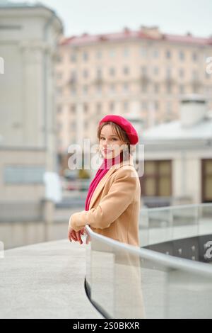 Eine Frau steht auf einem Balkon, warm gekleidet in einem beigen Mantel mit rosa Baskenmütze und Schal. Sie blickt nachdenklich über das Stadtbild hinaus und umschließt die Stockfoto