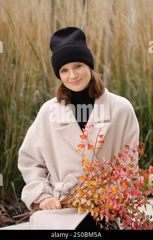 Eine fröhliche Frau sitzt friedlich umgeben von hohen Gräsern und trägt einen weichen beigefarbenen Mantel und eine schwarze Mütze. Stockfoto