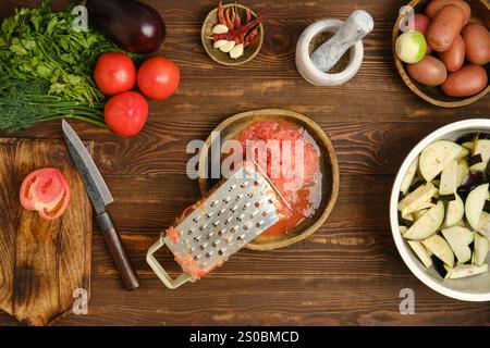 Geriebene frische Tomaten und Metallreibe auf einer Holzschale, umgeben von Auberginen, grünen Kräutern und Kartoffeln, sind auf einem Holztisch angeordnet. Stockfoto