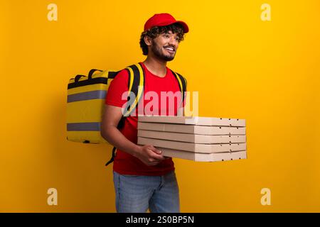 Ein junger Pizzen-Pizzen-Fahrer lächelt vor gelbem Hintergrund, trägt eine rote Mütze und ein rotes Hemd und strahlt lebendige Energie aus. Stockfoto