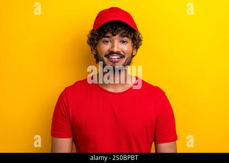 Stilvoller junger Mann in rotem Hemd und Mütze, der vor einem leuchtend gelben Hintergrund lächelt und Selbstvertrauen und Glück zeigt Stockfoto