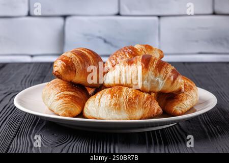 Frisch gebacken im Ofen knusprige und buttrige Croissants auf einem Teller auf schwarzem Holztisch mit Backsteinwand im Hintergrund, horizontale Ansicht, Nahaufnahme Stockfoto