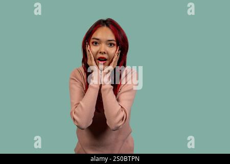 Studio-Aufnahme einer überraschten jungen Frau mit gefärbten roten Haaren, die das Gesicht mit den Händen hält und Staunen zeigt Stockfoto