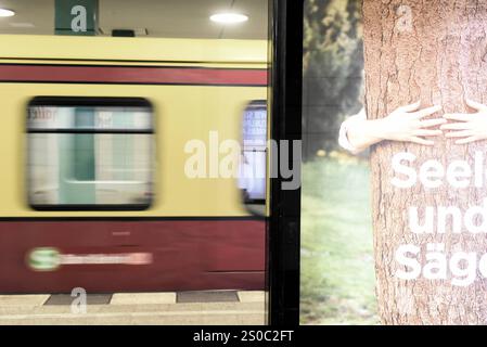 Auf einer Werbetafel am Bahnsteig S-Bahn ist ein Baum zu sehen, der umarmt wird und der Schriftzug Seele und Säge teilweise abgebildet wird. IMH Hintergrund fährt bewegungsunscharf eine S-Bahn ein. Werbung am Bahnsteig Berlin Anhalter Bahnhof *** auf einer Werbetafel auf dem Bahnsteig der S-Bahn ist ein Baum umfaßt und die Schrift Seele und Säge teilweise im Hintergrund abgebildet, eine S-Bahn kommt unbeweglich an Werbung auf dem Bahnsteig Berlin Anhalter Bahnhof Stockfoto