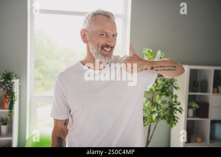 Lächelnder, grauhaariger älterer Mann, der zu Hause in einem hellen Raum die Daumen nach oben gibt und einen gesunden Lebensstil und eine positive Einstellung verkörpert Stockfoto