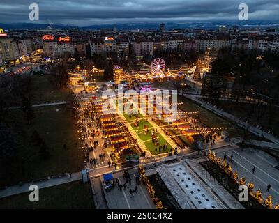 Ein fesselnder Blick aus der Vogelperspektive auf Sofias Weihnachtsmarkt in der Abenddämmerung in der Nähe von NDK, mit leuchtenden Lichtern, festlichen Ständen, einem Riesenrad, und Feiertagsstimmung. Stockfoto