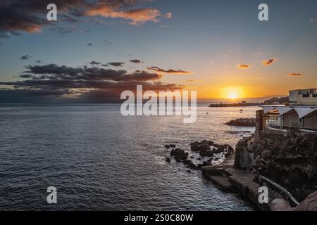 Wunderschöner Sonnenuntergang über einem ruhigen Meer mit felsiger Küste und bezaubernden Küstengebäuden, die eine ruhige und malerische Atmosphäre schaffen. Stockfoto