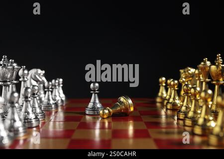 Silberner Bauer in der Nähe von gefallenem goldenem und anderen Schachfiguren auf Schachbrett vor schwarzem Hintergrund, Nahaufnahme. Wettbewerbskonzept Stockfoto