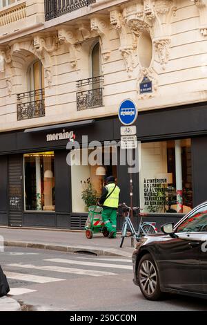 Ein Arbeiter schiebt während der Tageslichtstunden einen Wagen auf der belebten Straße vor einem modernen Geschäft in Paris Stockfoto