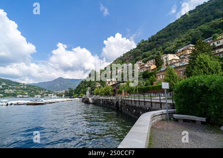 Eine malerische Uferpromenade entlang eines Sees, umgeben von üppigen grünen Hügeln und eleganten Villen am Hügel unter einem hellblauen Himmel mit flauschigen weißen Wolken Stockfoto