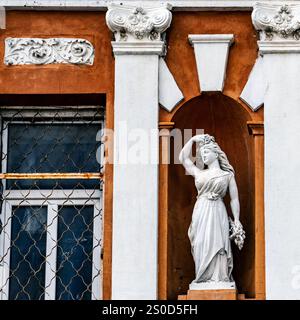 Eine klassische weiße Statue einer Frau mit Blumen steht in einer Nische an einer Gebäudefassade. Die Architektur verfügt über kunstvolle Details und ein Fenster wi Stockfoto