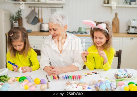 Frohe Ostern. Zwei kleine Schwestern Zwillinge, Großmutter, die Eier malt und die gemeinsame Zeit genießt. Glückliche Familie Großmutter Enkelinnen Kinder bereiten sich auf Ostern vor. Christliche Festivaltradition des Frühlings Stockfoto