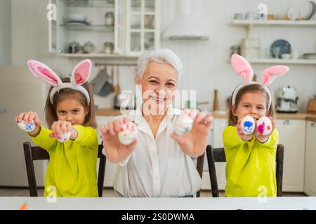 Frohe Ostern. Zwei kleine Schwestern Zwillinge, Großmutter, die Eier malt und die gemeinsame Zeit genießt. Glückliche Familie Großmutter Enkelinnen Kinder bereiten sich auf Ostern vor. Christliche Festivaltradition des Frühlings Stockfoto