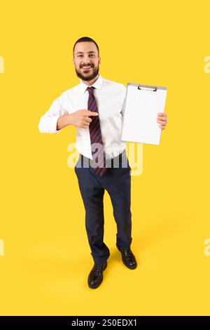 Junger Geschäftsmann, der auf leere Zwischenablage auf gelbem Hintergrund zeigt Stockfoto