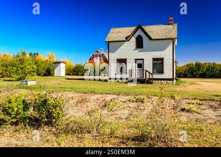 Ein kleines Haus mit weißem Dach sitzt auf einem Feld. Das Haus ist von Bäumen und einer Scheune umgeben. Der Himmel ist klar und blau Stockfoto