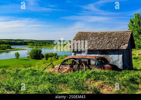 Ein verrostetes altes Auto parkt vor einem kleinen blauen Haus. Das Auto ist von Gras umgeben und das Haus liegt auf einem Hügel mit Blick auf einen Fluss Stockfoto