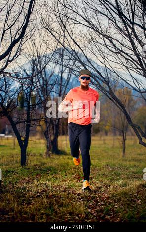 Sportlicher bärtiger Mann in rotem Hemd, der bei Sonnenaufgang im Herbstwald auf dem Wanderweg läuft Stockfoto